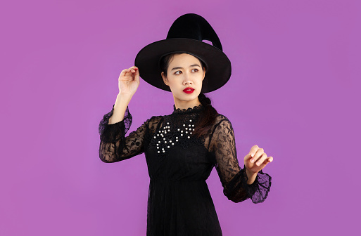 Young beautiful asian woman in black dress wearing witch hat in a concept of halloween costume posing on purple background.