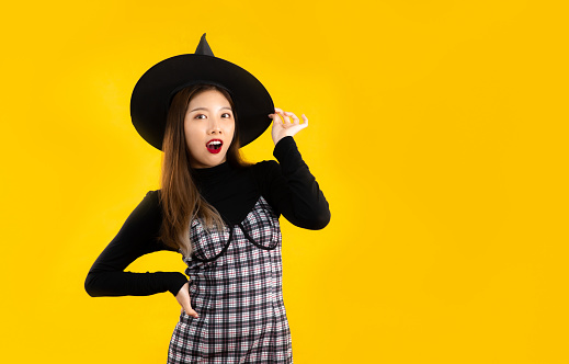 Young asian woman in halloween costume theme wearing witch hat posing on yellow background.