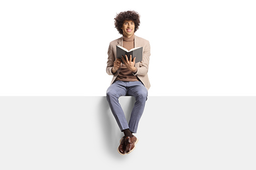 Elegant young man with curly hair sitting on a panel and holding a book isolated on white background