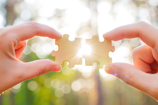 Hands put together wooden jigsaw puzzles against a bokeh forest background with sunlight coming through. Creative partnership concept.