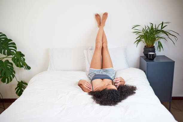 une jeune femme multiethnique se détend dans sa chambre de garde les jambes contre le mur - sleeping lying on back couple bed photos et images de collection