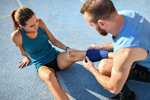 Male Coach Helping Female Athlete To Bandage Knee After Injury