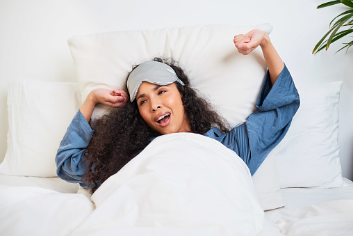 A young multi-ethnic woman stretches with arms behind head and sleep mask. High quality photo