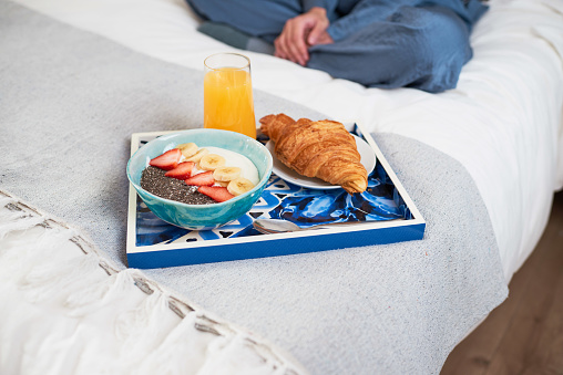 Close up of breakfast tray with croissant, fruit bowl on bed with woman sitting. High quality photo