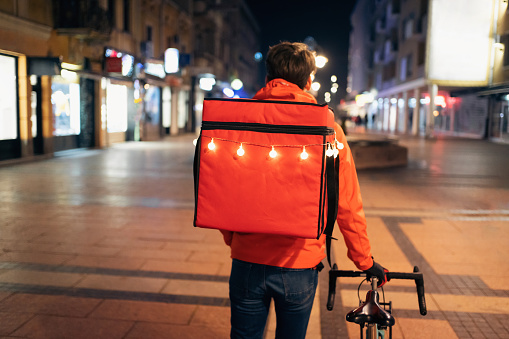 Rear-view of unrecognizable male delivery person, pushing bicycle through city, while having delivery bag decorated with Christmas lights