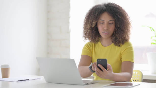 istock African Woman Browsing Internet on Smartphone while using Laptop in Office 1423322976