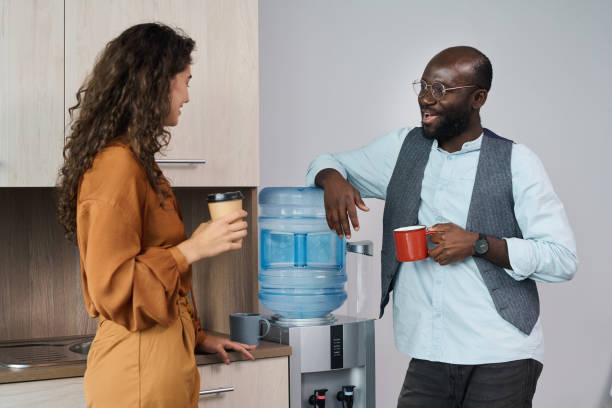 Happy young intercultural office workers having tea and coffee at break Happy young intercultural office workers chatting and having tea and coffee at break while standing by water dispenser with gallon cooler stock pictures, royalty-free photos & images