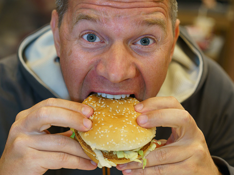 Portrait of hungry man aged 40-50 y.o. eating fast food in city cafe. He eagerly bites off a piece of burger and looks at camera. He stared. Defocused cafe interior as background.