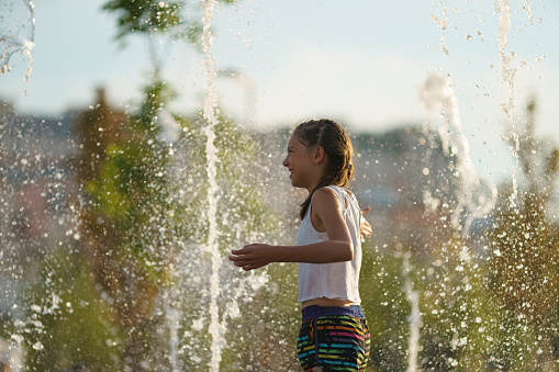 Moscow, Russia - June 21, 2021: Happy beautiful girl touching water of fountain sprays. She is happy and smiling. Hot summer day. Time for refreshment. Recreation concept.