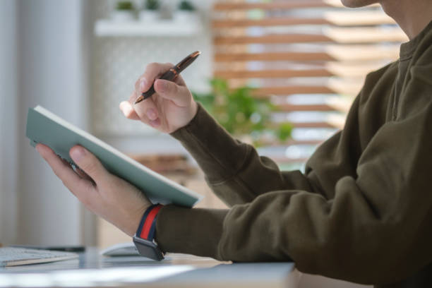 istock Cropped view of young man sitting at home office desk and making notes on notebook. 1423339200