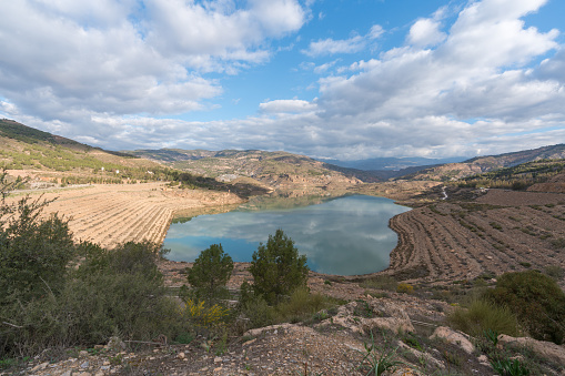 Beninar reservoir in the south of Spain, there are pine trees and rocks, the sky has clouds