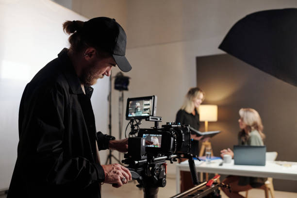 Young serious production studio worker adjusting videocamera during shooting Young serious production studio worker adjusting videocamera during shooting of commercial with participation of two women behind the scenes stock pictures, royalty-free photos & images