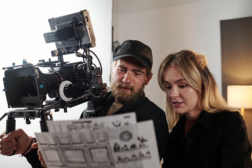 Bearded videographer with videocamers and his blond female assistant discussing working documents during studio shooting