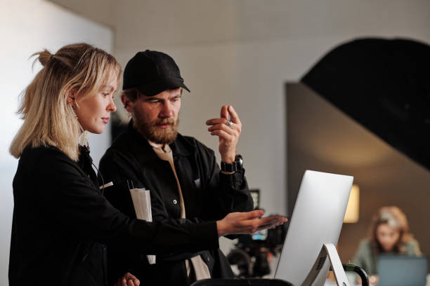 Young female assistant of videographer pointing at computer screen Young female assistant of videographer pointing at computer screen during discussion of new commercial in production studio film-crew stock pictures, royalty-free photos & images