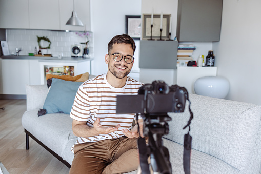 Young smiling man vlogging and talking for his audience. He is sitting on the sofa in the living room and talking in front camera
