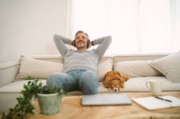 pria kaukasia usia paruh baya yang tenang duduk di sofa mendengarkan musik menikmati meditasi untuk tidur dan pikiran yang damai di headphone nirkabel, bersandar dengan anjing chihuahua-nya yang cantik duduk di sampingnya. - kehidupan domestik subjek potret stok, foto, & gambar bebas royalti