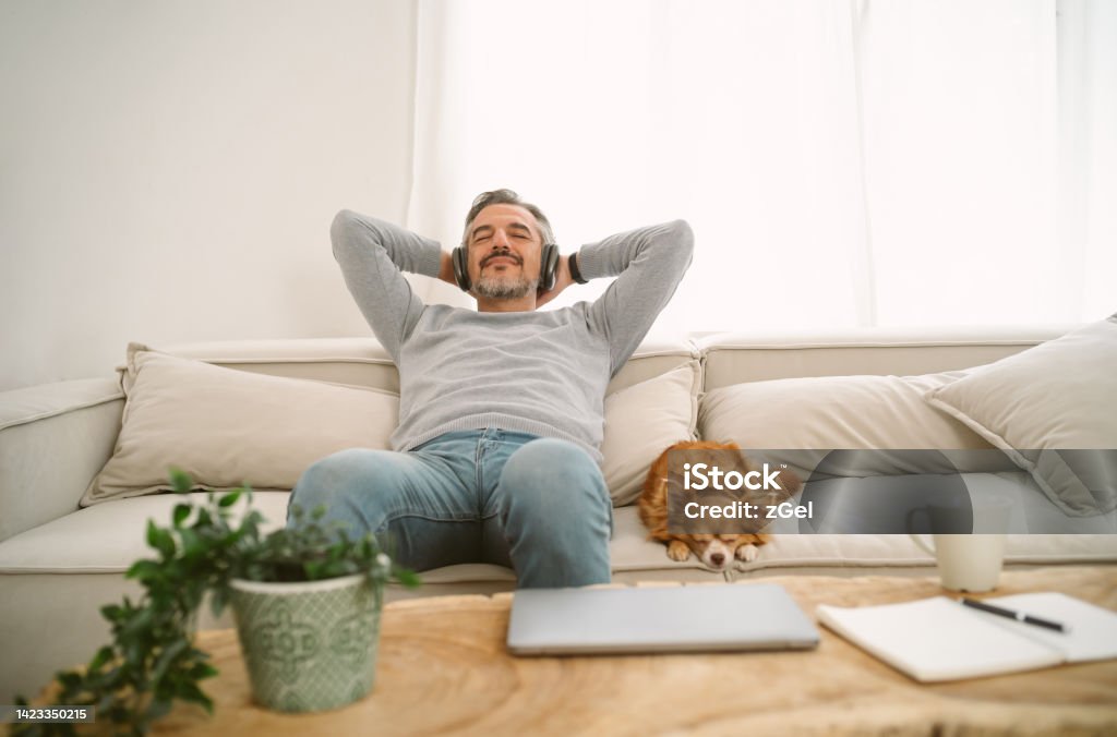 Calm Middle age Caucasian man sitting on sofa listening to music enjoying meditation for sleep and peaceful mind in wireless headphones, leaning back with his lovely chihuahua dog sit besides. Relaxation Stock Photo