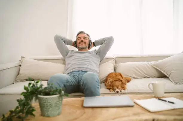 Photo of Calm Middle age Caucasian man sitting on sofa listening to music enjoying meditation for sleep and peaceful mind in wireless headphones, leaning back with his lovely chihuahua dog sit besides.