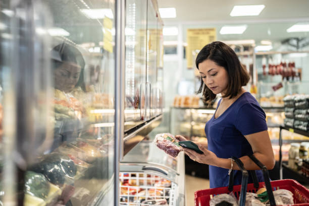 donna adulta di metà che usa il telefono cellulare che compra generi alimentari in un supermercato - meat supermarket butchers shop market foto e immagini stock