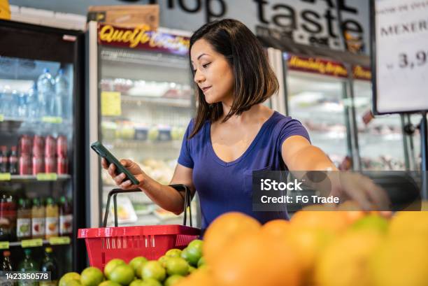 Mid Adult Woman Using The Mobile Phone While Choosing Orange In A Supermarket Stock Photo - Download Image Now