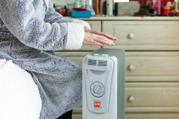 Senior woman warming her hands over electric heater at home A retired senior woman in her 70s sits at home inside her cold house in winter. It is so cold that she is wrapped up in warm winter clothing, and is holding her hands over an electric heater for some extra warmth and comfort. Selective focus with room for copy space. electric heater stock pictures, royalty-free photos & images