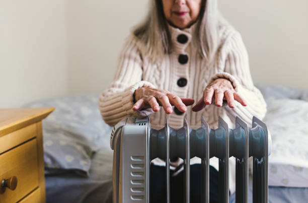 Senior woman warming her hands over electric heater at home A retired senior woman in her 70s sits at home inside her cold house in winter. It is so cold that she is wrapped up in warm winter clothing, and is holding her hands over an electric heater for some extra warmth and comfort. Selective focus with room for copy space. cost of living stock pictures, royalty-free photos & images