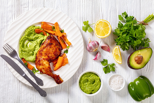 fried chicken leg with guasacaca sauce of avocado, green pepper and herbs and sweet potato chips on white plate on wooden table with ingredients, horizontal view from above, flat lay