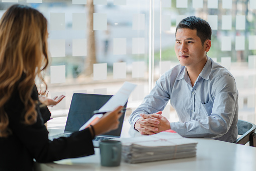 Business cooperation concept of two young Asian business partner organizations: man and woman sitting at desk with laptop working paperwork together. Colleagues discuss company finances.