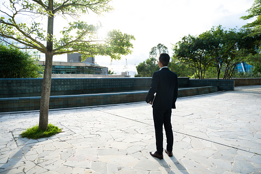 Back view of a young Asian businessman wearing formal suit looking out at cityscape in the outdoor workpace of a modern office, thinking about plans and future. Portrait of a confident Asian execitive staring away at city views planning his career, feeling hopeful.