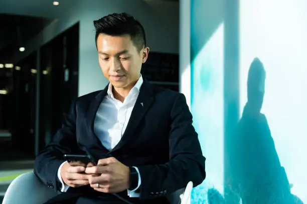 Young Asian businessman wearing formal suit sitting in a modern office space while holding a smartphone. Portrait of an Asian man in black suit sitting on a chair indoors in an office building, looking at the device screen of his mobile phone, texting people.