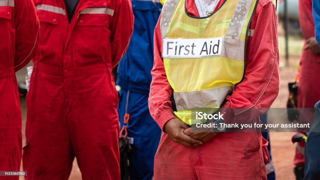 First Aid person in emergency drill. Group meeting of field operation staffs with a man who wear "First Aid" vest, preparing for emergency drill and practice scenario at construction site. Industrial people working scene photo. Evacuation Stock Photo