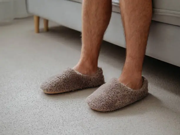 Cropped shot of unrecognizable man in soft plush warm slippers, sitting on sofa in morning time at cozy scandinavian home. Hairy legs of adult man in trendy warm slippers on floor