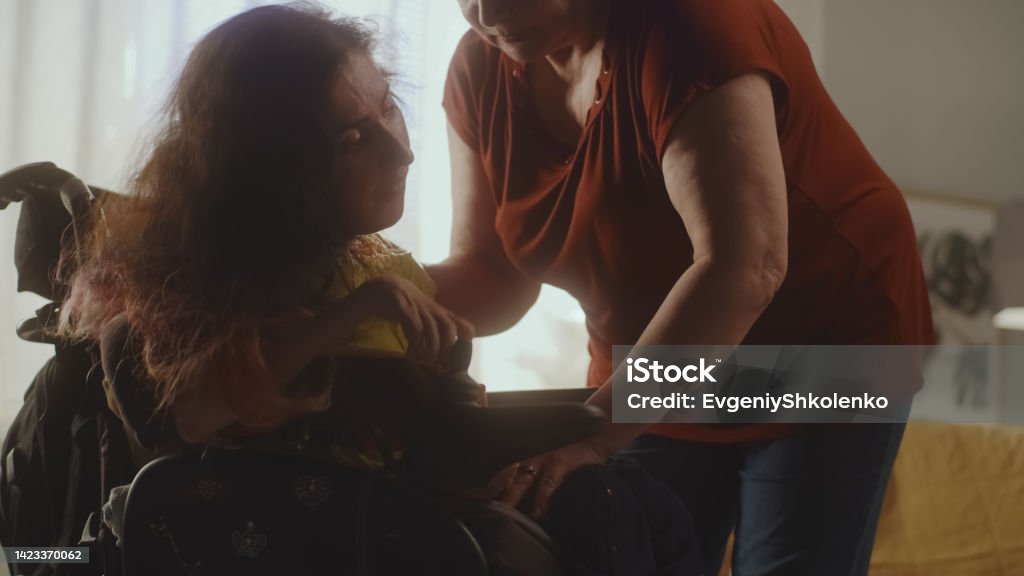 Caregiver helping woman to sit in a wheelchair Aged woman caregiver helping woman with a spinal muscular atrophy setting a modern motorized wheelchair for comfortable sitting Legal Guardian Stock Photo