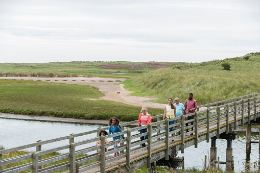 A wide-angle view of a family enjoying the coastal beach trail in Beadnell in the North East of England. The youngest members of the family are strides ahead and leading the way as they walk across a wooden walkway.