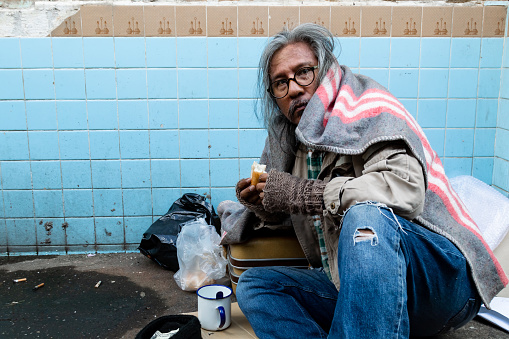 A homeless Asian old man with a long beard sits on the street eating bread after receiving a delicious snack. from the people passing by The poor have no homes on the streets.