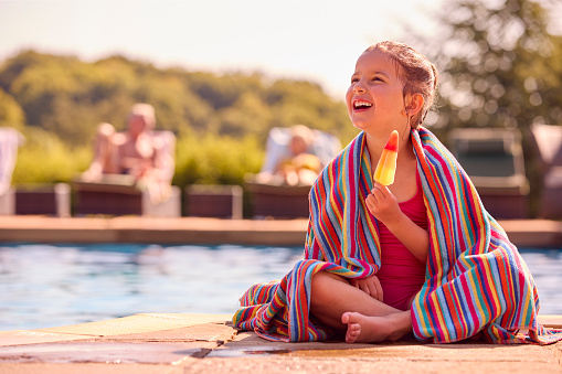 Family On Summer Holiday With Girl Wrapped In Towel Eating Ice Lolly By Swimming Pool