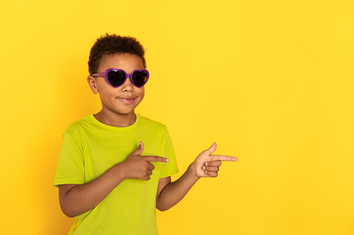 Portrait of happy preteen boy in sunglasses pointing direction. Mixed race child wearing green T-shirt showing something against yellow background. Presentation concept