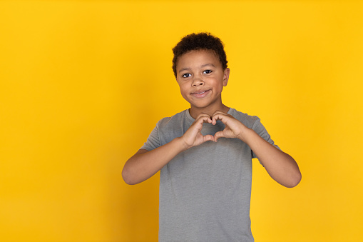 Portrait of happy preteen boy making heart gesture. Mixed race child wearing gray T-shirt showing love symbol, looking at camera and smiling at camera. Love or admiration concept