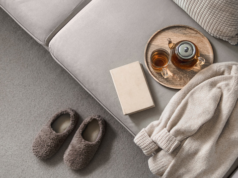 Cashmere sweater, reading and serving tray on gray sofa. Warm weekend at home aesthetics. Detail of cozy scandinavian winter interior. Warm soft winter slipper on carpet at home