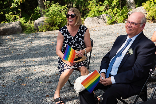 Parents of the bride attending LGBTQ+ wedding. They are holding the rainbow flag. This is part of a series about a lesbian couple getting married. Horizontal outdoors waist up shot with copy space.