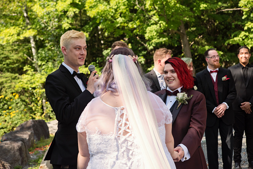 Lesbian brides at LGBTQ+ wedding ceremony outdoors in summer. Celebrant holding the mic, brides are holding hands. Grooms are in the background. This is part of a series about a lesbian couple getting married. Horizontal outdoors waist up shot with copy space.