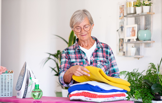 Caucasian smiling senior woman in checkered shirt ironing clothes at home on ironing board