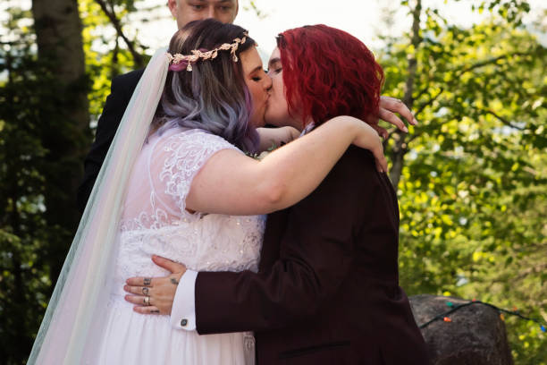Lesbian brides at LGBTQ+ wedding ceremony outdoors in summer. Lesbian brides at LGBTQ+ wedding ceremony outdoors in summer. Celebrant in the middle, newlywed brides kissing. This is part of a series about a lesbian couple getting married. Horizontal outdoors waist up shot with copy space. gay wedding kiss stock pictures, royalty-free photos & images