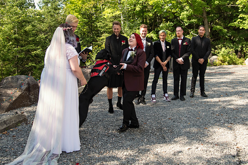 Lesbian brides at LGBTQ+ wedding ceremony outdoors in summer. Service dog is in action. This is part of a series about a lesbian couple getting married. Horizontal outdoors full length shot with copy space.