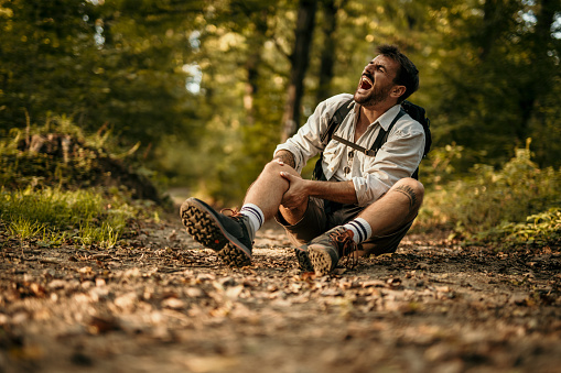 Cool looking nature lover, a hiker, felt down while hiking in nature with his backpack, screaming and holding his knees that hurts. Adventure is not always a good memory