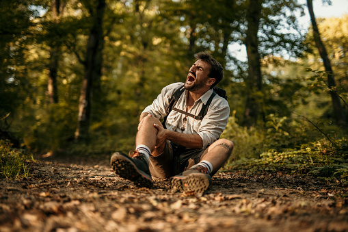 Tired caucasian man in huge pain with dislocated knee hiking alone in summer nature. Hiking, travel outdoor, recreation, and active adventure concept