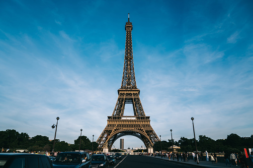 Cars riding on road and tourists walking on sidewalk near famous Eiffel Tower against cloudy blue sky in Paris, France