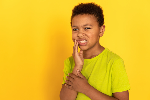 Portrait of sad preteen boy suffering from toothache. Mixed race child wearing green T-shirt touching aching cheek against yellow background. Childs health problems concept