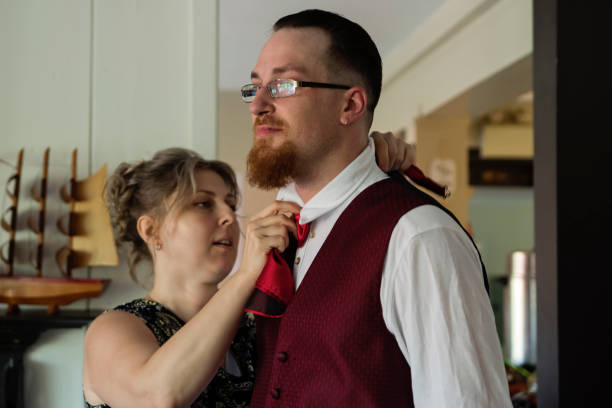 Friends getting ready for LGBTQ+ wedding. Woman helping trendy young man to put on a necktie. This is part of a series about a lesbian couple getting married. Horizontal indoors waist up shot with copy space.