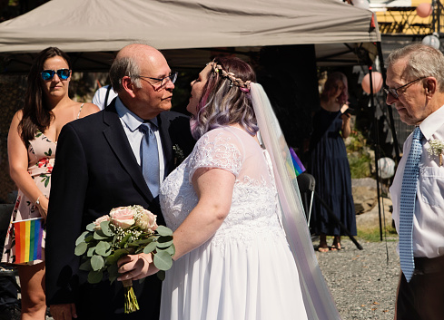 Father and grandfather walking daughter bride down the aisle for LGBTQ+ wedding. This is part of a series about a lesbian couple getting married. Horizontal outdoors mid waist shot with copy space.